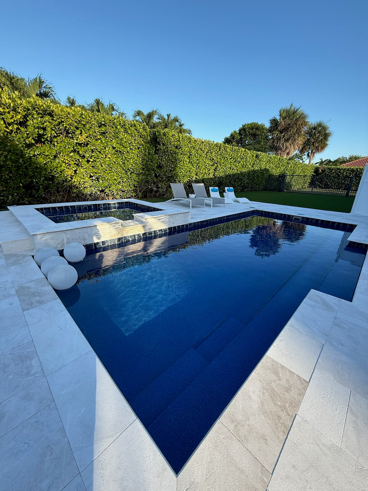 Rectangular fiberglass pool with raised spa, white travertine deck, loungers and hedge, installed by Cool Pool of Florida